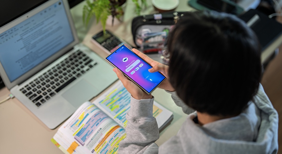 Et barn holder en smartphone med en lys appskærm over farvekodede noter ved siden af en åben bærbar computer. Foto: Getty Images.